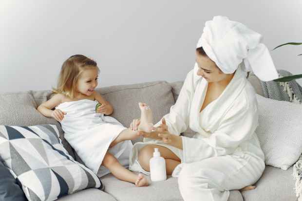 woman in white robe sitting on gray couch putting lotion on little girls foot